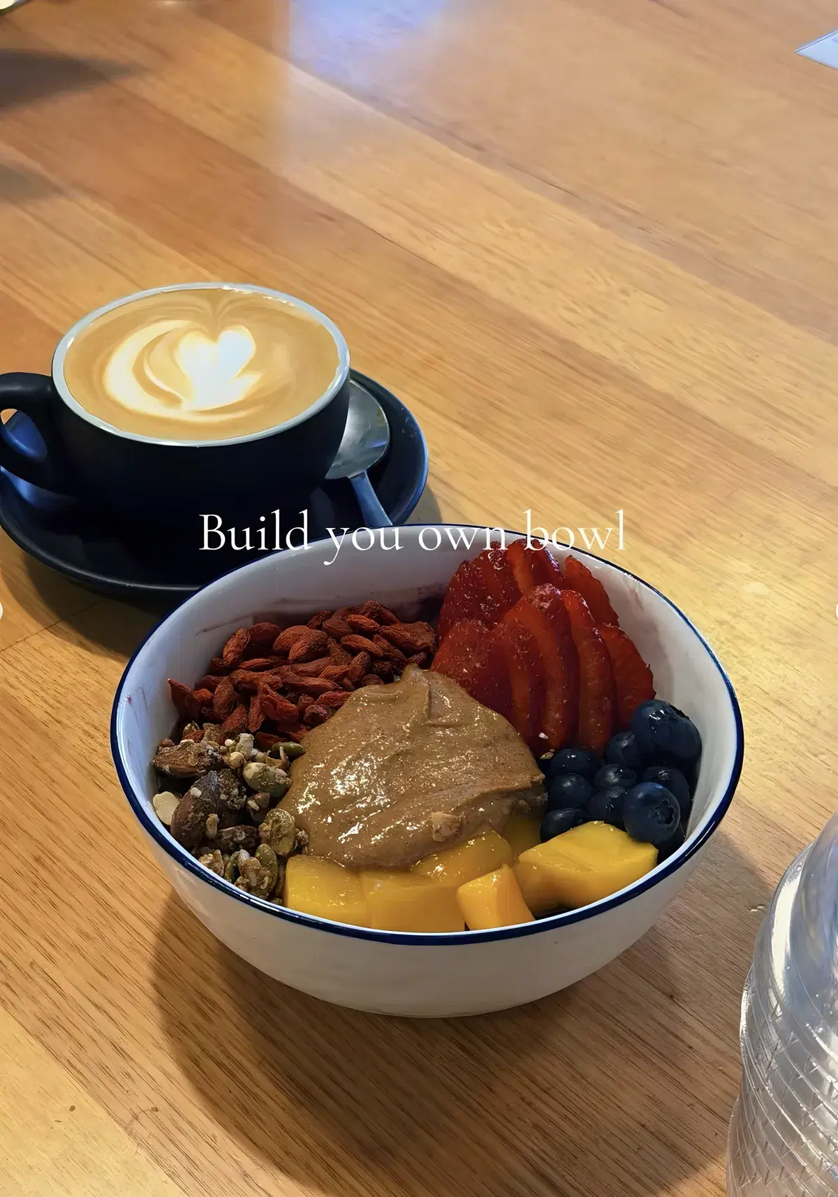 Build-your-own acai bowl with peanut butter, goji berries, granola, mango and berries, beside a latte